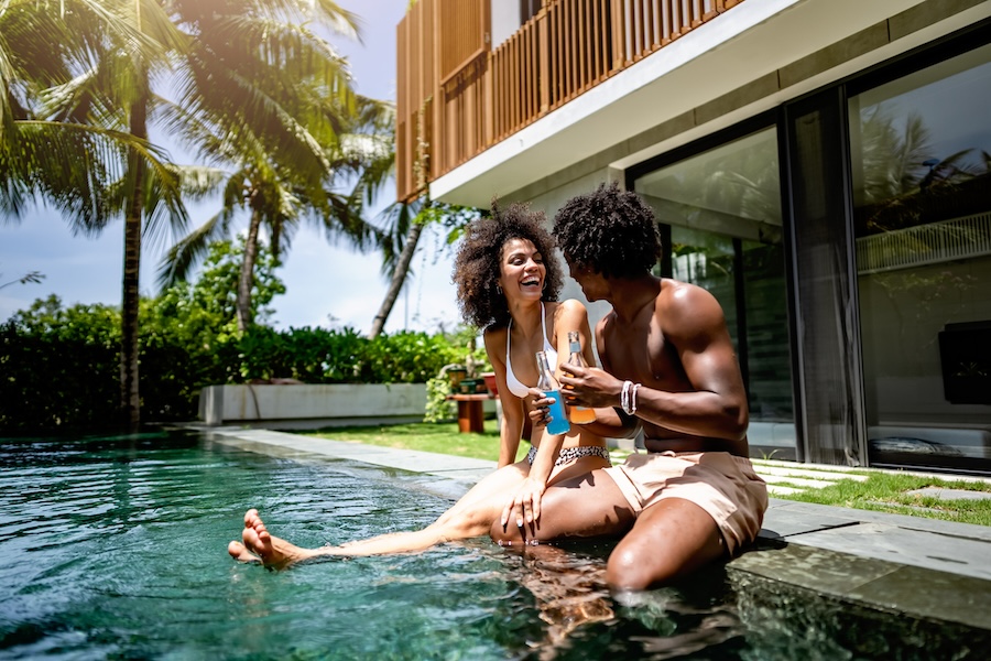 A young couple of African descent enjoying a sunny day at a luxurious poolside, sitting at the pool's edge with drinks in hand.
